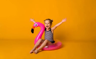 Çizgili mayo giymiş mutlu küçük sarışın kız flamingo ile pembe şişirilebilir bir çemberde oturuyor ve sarı bir arka planda mesaj için bir yere sahip kameraya bakıyor. Bir çocukla yaz tatili