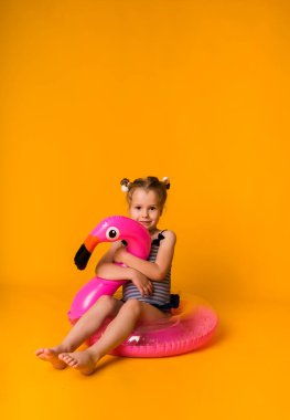 Çizgili mayo giymiş küçük bir kız pembe şişirilebilir bir dairede oturuyor ve sarı arka planda flamingo ve mesaj için yeri var. Bir çocukla yaz tatili