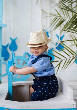 Hasır şapkalı küçük bir beyefendi ahşap bir kayıkta oturuyor ve arka planda deniz süslemeleri var.