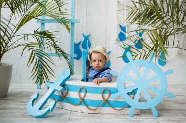 Takım elbiseli küçük bir gezgin ve hasır şapkalı arka planda mavi ve beyaz ahşap bir teknede oturuyor.