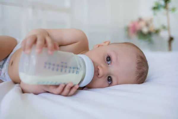 赤ん坊はベッドの上の綿の毛布の上に寝そべってボトルから混合物を食べる 人工給餌 — ストック写真