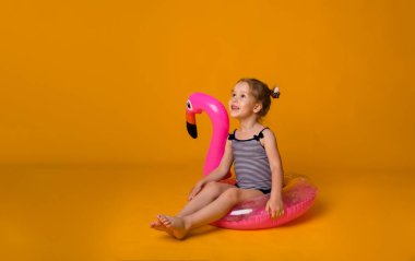 Çizgili mayo giymiş küçük sarışın kız flamingolu pembe şişirilebilir bir dairede oturuyor ve sarı bir arka plana bakıyor.