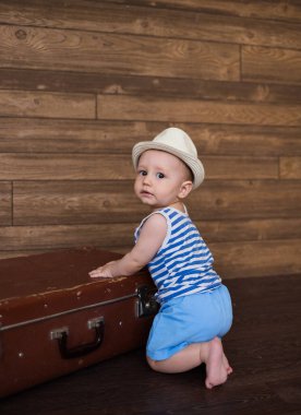 Çizgili tişörtlü ve şortlu küçük bir gezgin ahşap kahverengi bir arka planda eski bir bavulla oturur.