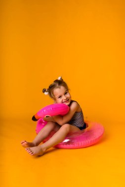 Çizgili mayo giymiş mutlu küçük sarışın kız pembe şişirilebilir bir çemberin içinde oturuyor ve sarı arka planda bir flamingoya sarılıyor.