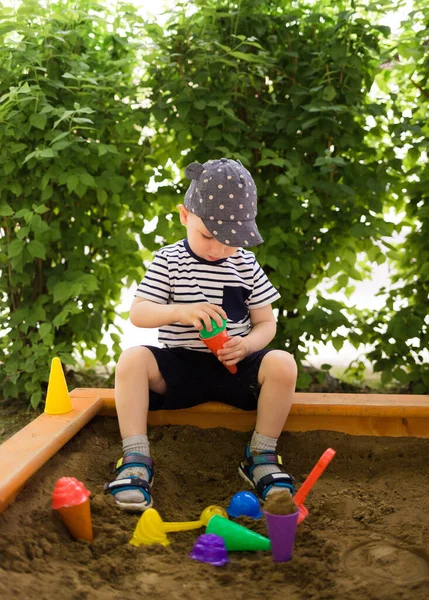 Şapkalı küçük çocuk kum havuzunda oturur ve spatula ve küflerle oynar. Yaz Oyunları