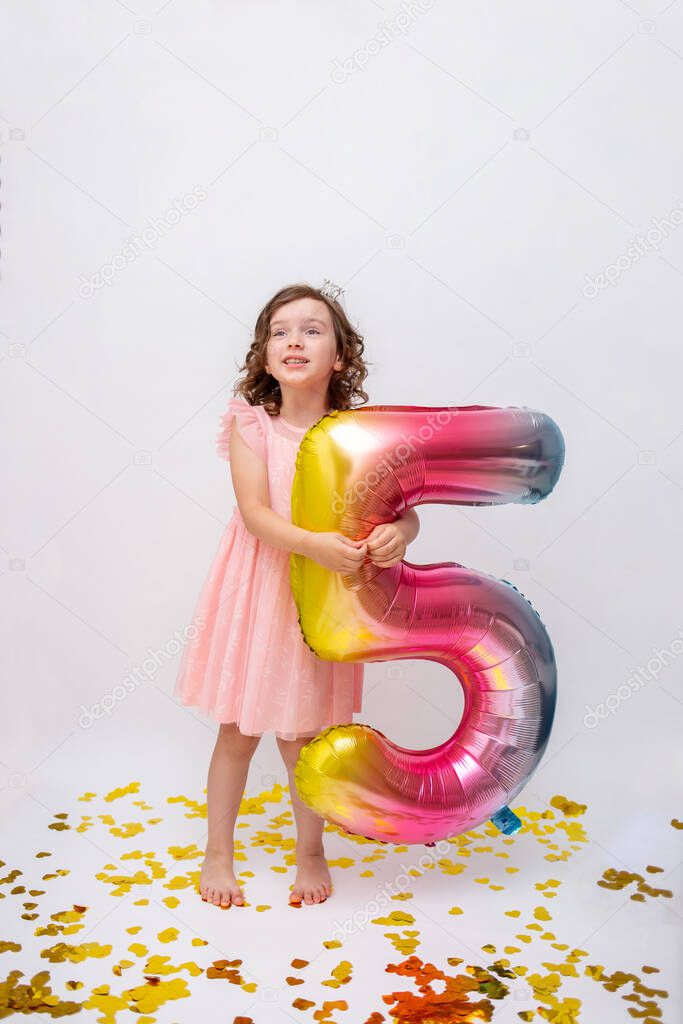 little curly-haired girl in a festive dress stands with an inflatable ball with the number five on a white background with gold confetti. Birthday