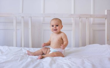 Bebek bezi takmış beyaz pamuklu bir yatakta beyaz bir kız oturuyor ve kameraya bakıyor.