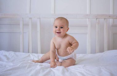 Bebek bezli mavi gözlü kız odadaki beyaz pamuklu battaniyenin üzerinde oturuyor.