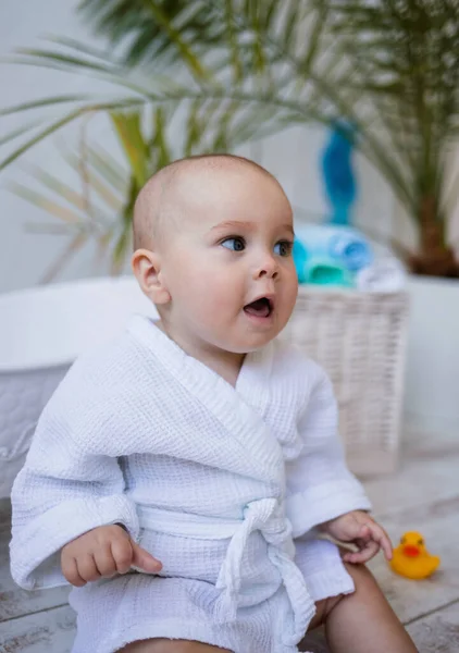 surprised baby girl in a white robe is sitting near a bath on a white background with a place for text. Water treatments for children