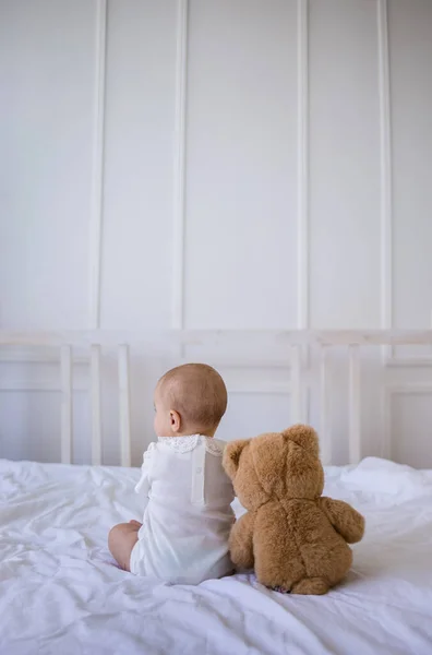 baby girl in a white bodysuit is sitting with her back with a teddy bear toy on a white cotton blanket on the bed in the room