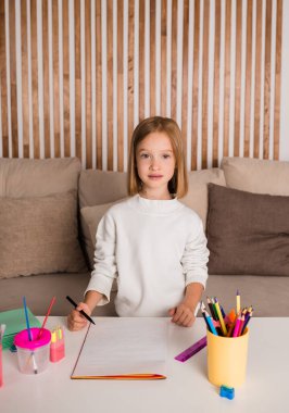 Küçük sarışın bir kız masada oturuyor ve kağıda resim çiziyor. Çocuklar için sanat