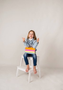 Kot pantolon giymiş mutlu, küçük bir sarışın kız beyaz bir arka planda ders kitaplarıyla oturuyor ve mesaj için bir yeri var. Okula geri dön.