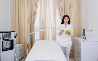 Beyaz üniformalı bir kozmetikçi ofiste duruyor ve ürünü yeşil bir kasede karıştırıyor.