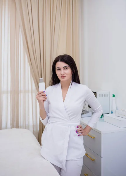 Beyaz üniformalı bir güzellik uzmanı ofiste ultrasonik yüz temizleme için bir cihaz tutuyor.