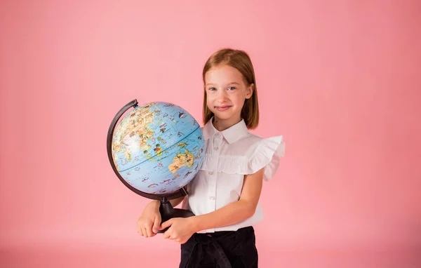 a blonde schoolgirl in a school uniform holds a globe on a pink background with a place for text