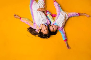 Pofuduk pijamalı mutlu küçük kızlar sarı arka planda uzayın bir kopyasıyla yatıyorlar.