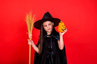 Cadı kostümlü, şapkalı küçük bir kız elinde süpürge ve kırmızı arka planda mesaj atacak bir yer olan bir balkabağı tutuyor.