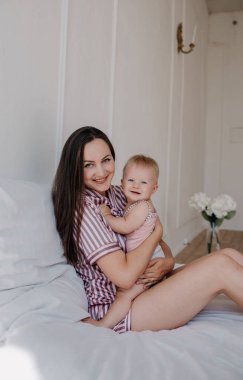 Pijamalı güzel esmer anne küçük kızını yatakta kucaklıyor ve kameraya bakıyor.