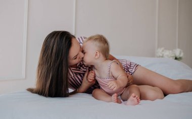 Çizgili pijamalı esmer bir anne yatağa uzanıp küçük kızını öper.