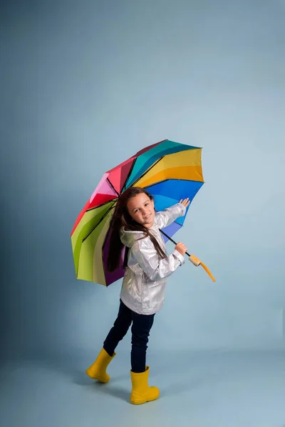 a little girl in a raincoat and yellow rubber boots holds a multi-colored umbrella on a blue background with a copy of the space