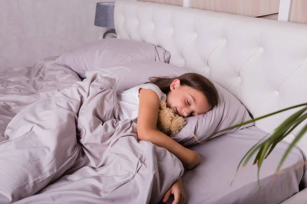 Pijamalı esmer bir kız yatak odasındaki yatakta yumuşak bir oyuncakla uyuyor.