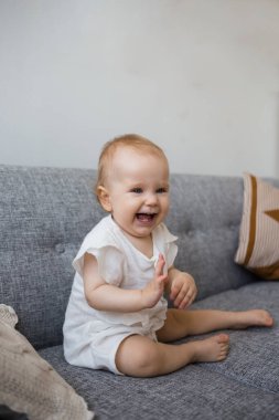 Muslin tulumlu gülen bir kız bebek oturma odasındaki kanepede oturuyor.