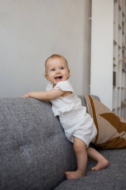 Muslin tulumlu mutlu bir bebek odada gri bir kanepede duruyor ve gülümsüyor.