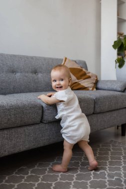 Muslin tulumlu bir bebek odada gri bir kanepenin yanında duruyor ve gülümsüyor.