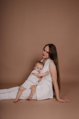 Beyaz takım elbiseli şık bir anne, bej arka planda, mesaj atabileceği bir yerde oturan bir bebekle oturuyor.