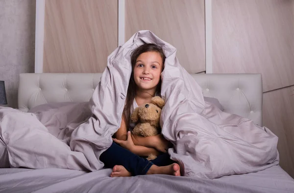 Pijamalı neşeli küçük bir kız yatakta battaniyenin altında oyuncak bir ayıyla oturuyor.