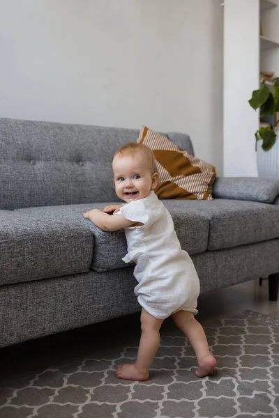Muslin tulumlu bir bebek odada gri bir kanepenin yanında duruyor ve gülümsüyor.