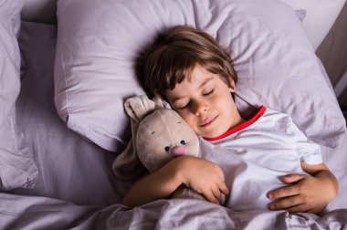 Pijamalı küçük bir çocuk pamuk bir yatakta içi doldurulmuş bir oyuncakla uyur.