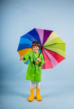 Yeşil yağmurluk ve sarı lastik çizmeli mutlu küçük bir çocuk mavi arka planda, üzerinde uzay kopyası olan çok renkli bir şemsiye tutuyor.