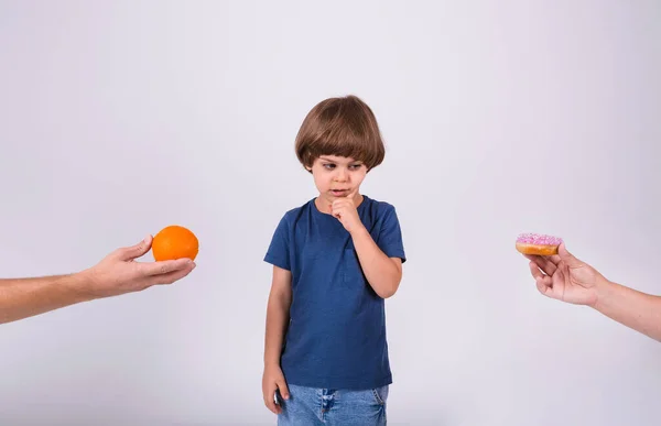 a little boy in a T-shirt makes a choice between an orange and a donut on a white background with a place for text