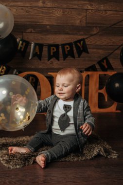Gri takım elbiseli bir doğum günü çocuğu kahverengi arka planda bir balonla oturuyor ve oynuyor.