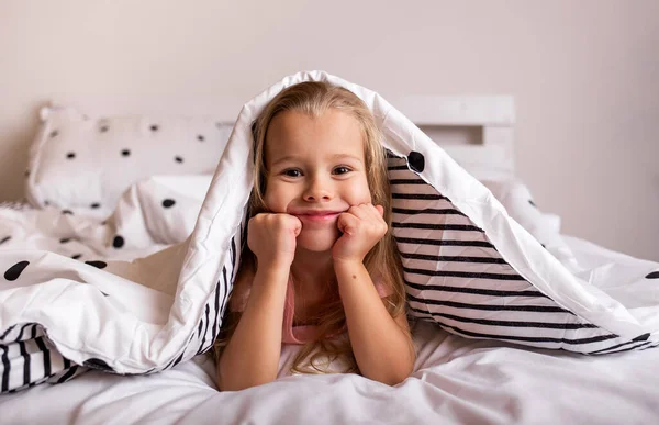 Küçük sarışın bir kız battaniyenin altında ve odada yatağında yatıyor.