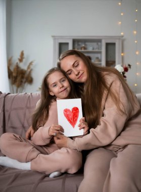 Güzel bir anne ve kız kanepede oturmuş, ellerinde bir kartpostalla kameraya bakıyorlar.