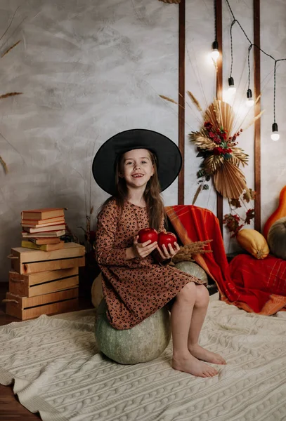 Cadı şapkalı küçük bir kız odada büyük bir balkabağının üzerinde oturuyor.