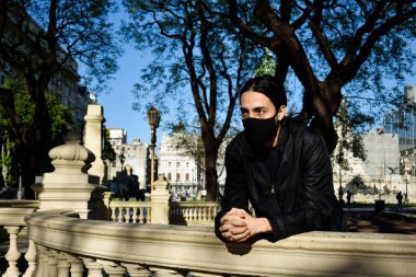 Avrupalı genç adam elinde maskeyle balkona bakıyor.