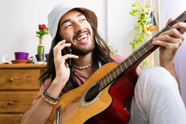 young hipster guitarist talking on the phone, with an acoustic guitar in his hands, composing on the phone, smiling, wearing a bucket hat
