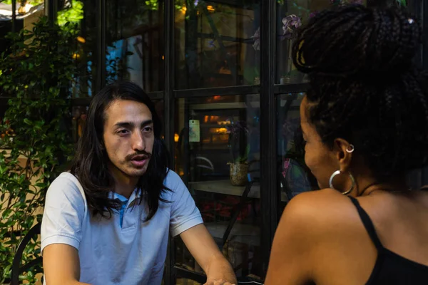 Young man with long hair on a date with a colored woman with braids. chatting among friends in a cafeteria with lots of nature, interracial place.