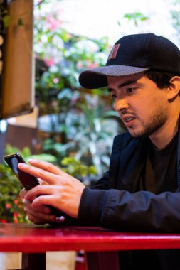 Elinde cep telefonu olan şapkalı genç bir adam açık hava restoranında kırmızı bir masada yemek bekliyor. Dikey fotoğraf