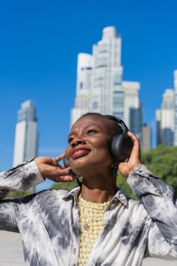 Siyah bir Afro-Amerikalı kadın, kablosuz kulaklıklı, şehir parkında gülümserken müzik dinliyor..