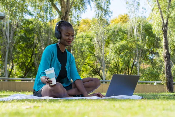 black african woman sitting on a blanket in the park using the computer and a wireless headset, while holding a hot drink