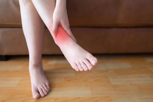 Woman suffering from ankle pain and sitting on sofa at home. Causes of pain include arthritis, tendinitis, diabetic neuropathy, gout or overuse injuries. Health care concept. Close up.