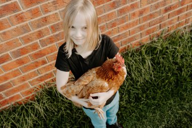 Genç sarışın kız elinde tavukla kameraya bakıyor.