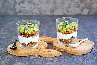 Ev yapımı yulaf ezmesi, fındık, kurutulmuş kızılcık ve bal ve şekersiz yoğurt ve koyu beton arka planda cam bir bardakta kivi. Sağlıklı kahvaltı..