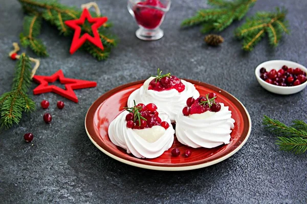 Portion Pavlova meringue pastries with cream and cranberry sauce in New Year\'s or Christmas decor on a red plate on a dark concrete background. New Year and Christmas desserts. Meringue recipes