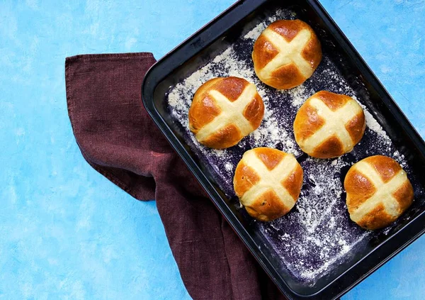 Traditional Easter cross buns made from yeast dough in a metal baking dish on a light blue concrete background. Easter holiday, concept. Easter recipes. British food