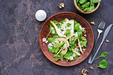 Taze ıspanak, sulu armut, ceviz ve parmesan salatası koyu beton arka planda kahverengi kil tabakta. Vejetaryen tarifleri. Sağlıklı yiyecekler.